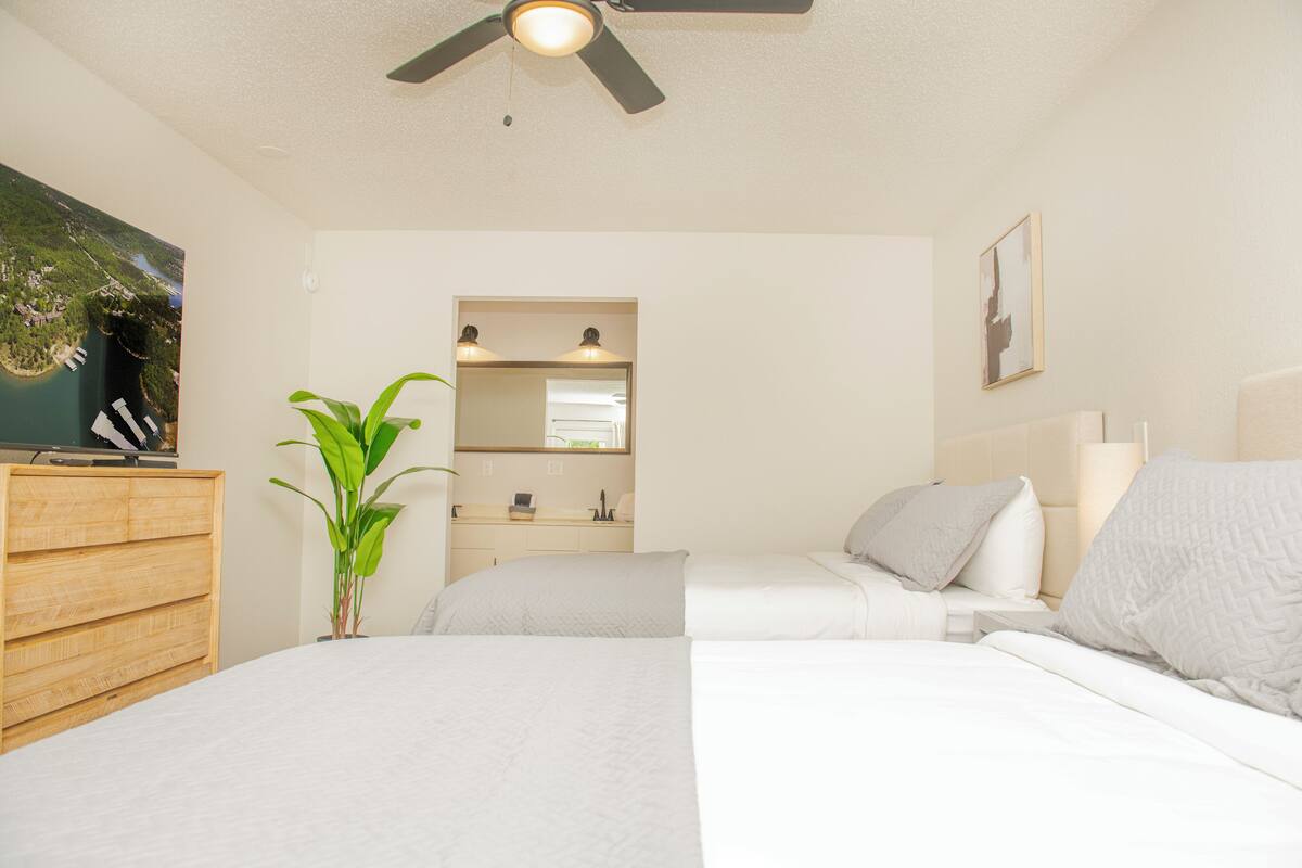 Une chambre d’hôtel avec deux lits, un ventilateur de plafond, une télévision, une plante, un miroir et un tableau au mur.