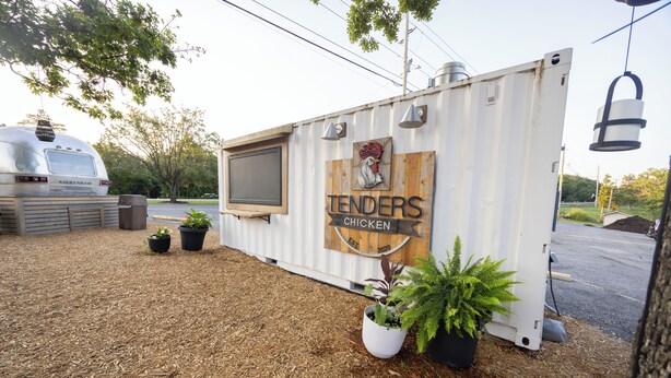 Un camión de comida móvil con un letrero que dice 'Tenders Chicken' y un logo de gallo, rodeado de mantillo y plantas en macetas.
