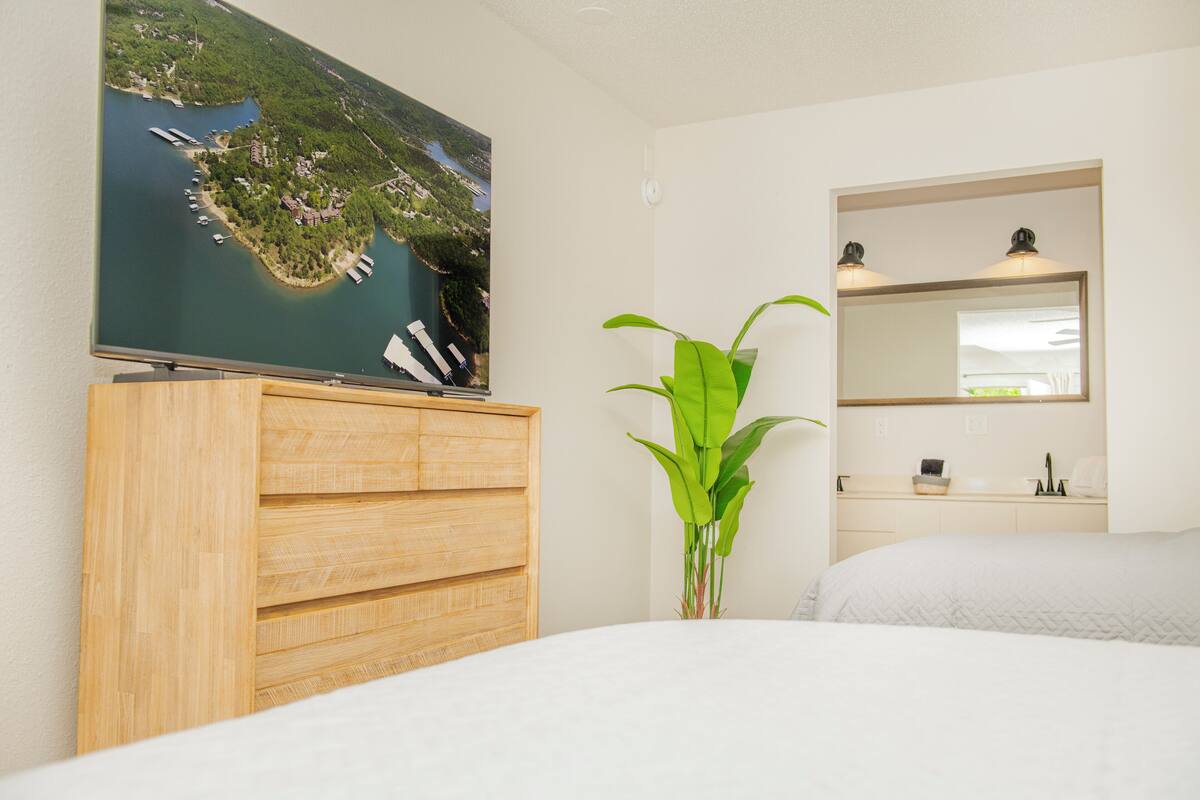 A hotel room with a bed, a wooden dresser, a large flat-screen TV mounted on the wall, a potted plant, a mirror, and a window with a view of a body of water.