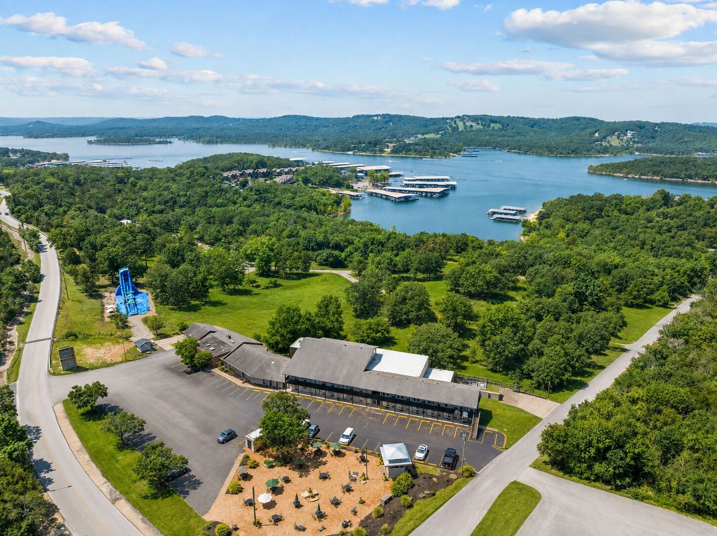 A hotel with a parking lot, a building with a flat roof, and a body of water with boats in the background.