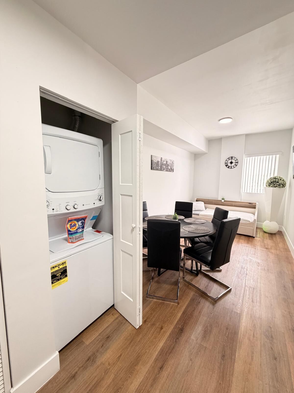 Une chambre moderne équipée d’un lave-linge et d’un sèche-linge intégrés, d’un coin repas avec une table ronde et des chaises, d’un lit avec une tête de lit et d’une horloge murale.
