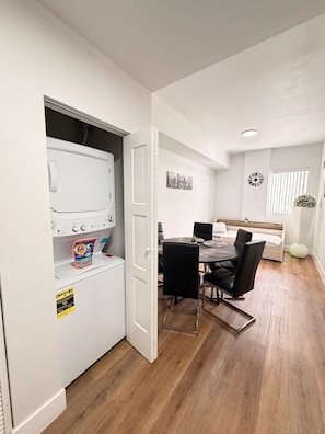 Ein modernes Zimmer mit integrierter Waschmaschine und Trockner, einem Essbereich mit rundem Tisch und Stühlen, einem Bett mit Kopfteil und einer Wanduhr.
