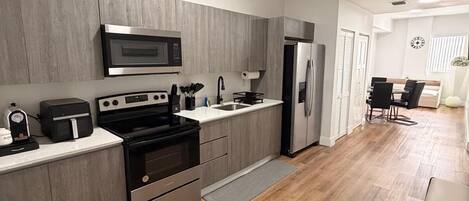 A modern kitchen with a microwave, oven, and refrigerator, a dining area with a table and chairs, and a hallway leading to another room.