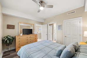 Ein Schlafzimmer mit Bett, Kommode, Fernseher, Deckenventilator und einer Tür zu einem anderen Raum.