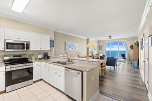 A compact kitchen with a microwave, oven, and dishwasher. There is a dining area with a table and chairs, and a view of the ocean through large windows. - High-Floor Gulf View & Balcony - Emerald Skies (Panama City Beach)