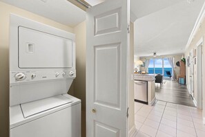 Eine Waschküche mit Waschmaschine und Trockner, ein Flur, der zu einem Zimmer mit Meerblick führt, und eine Tür mit einer goldfarbenen Klinke.