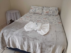 Ein ordentlich bezogenes Bett mit einem blumigen Überwurf, ein gefaltetes weißes Handtuch und ein kleiner weißer Tisch daneben.
