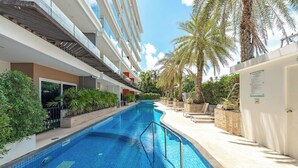 Una piscina con plataforma de baldosas, rodeada de palmeras y un edificio de varios pisos.