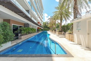 A swimming pool with a tiled deck, surrounded by palm trees and a multi-story building. - Luxury Babylon Sky Pool & Ocean View C189 (Tambon Rawai)