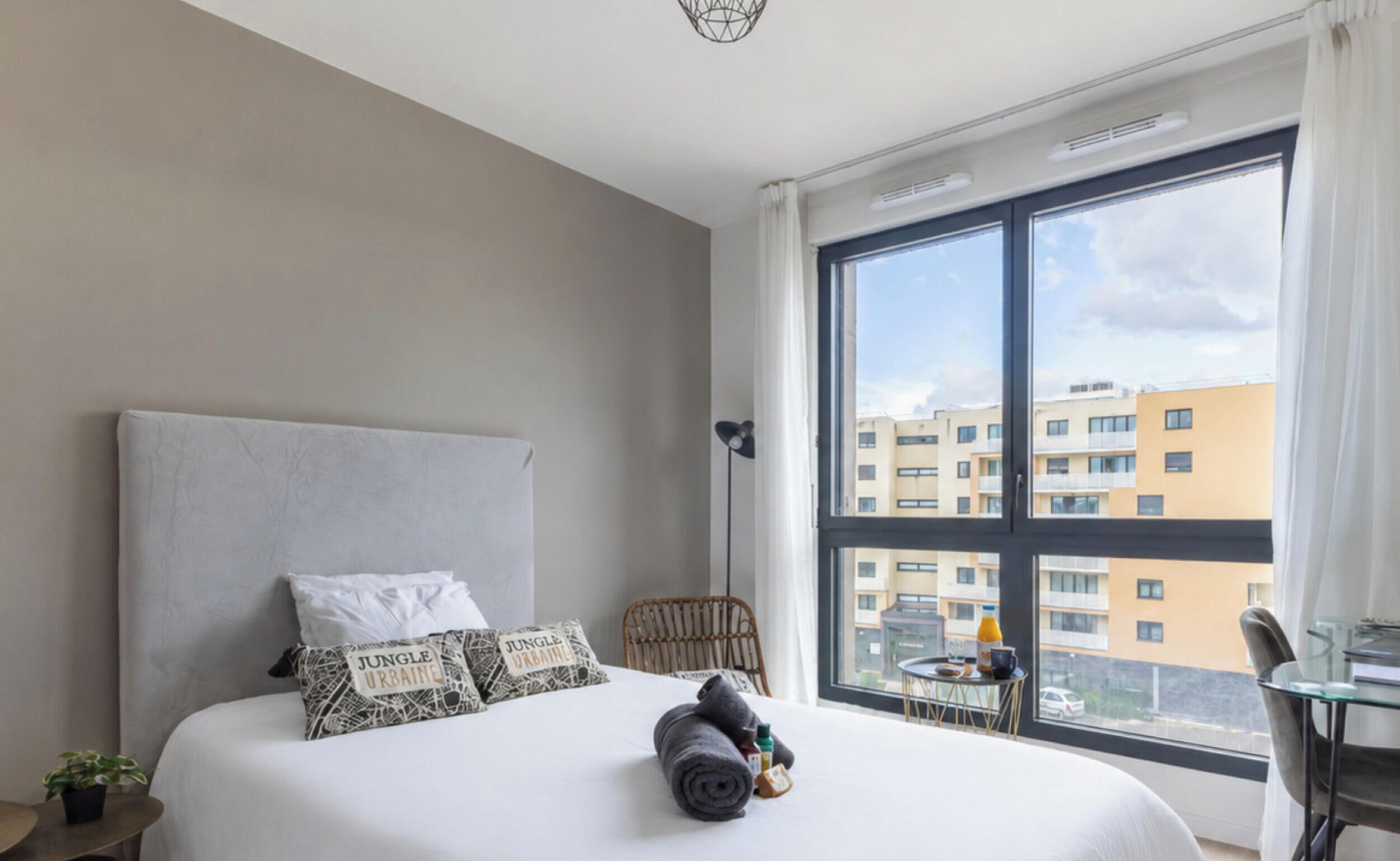 Habitación de hotel con una cama grande, vistas a edificios, una mesita con bandeja y una silla.