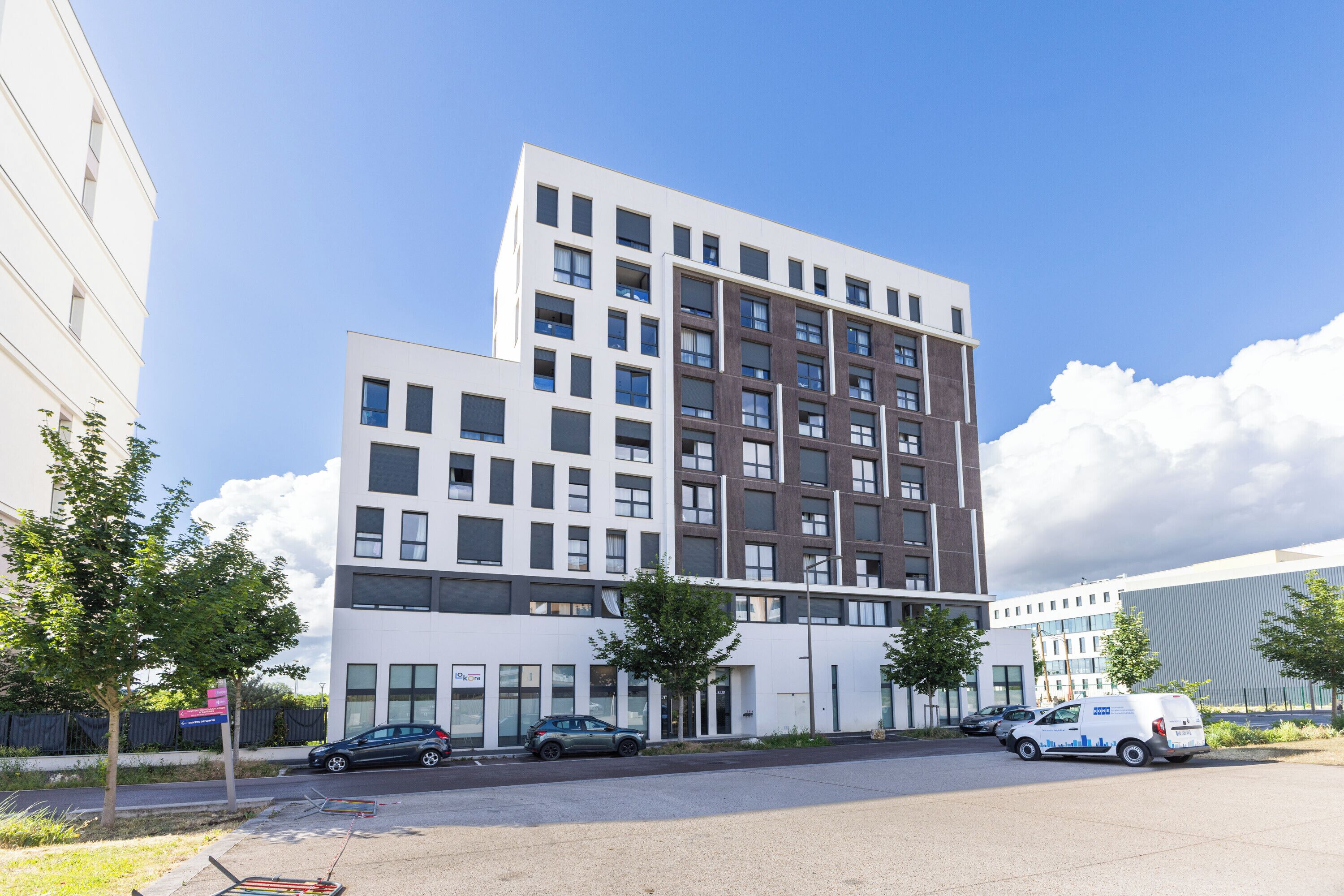 A modern multi-story building with a flat roof, large windows, and a parking area with cars.