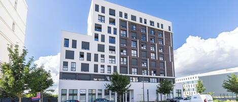 A modern multi-story building with a flat roof, large windows, and a parking area with cars.