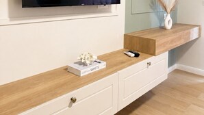Una sala de estar moderna con un televisor de pantalla plana montado en la pared, una consola de madera con cajones, un pequeño jarrón con flores secas y un espejo redondo sobre una pared de color claro.