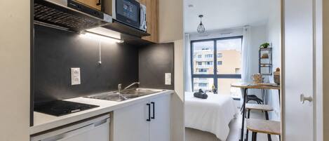 A compact kitchen with a microwave, stove, and sink. There is a small bar area with stools and a window with a view of buildings.