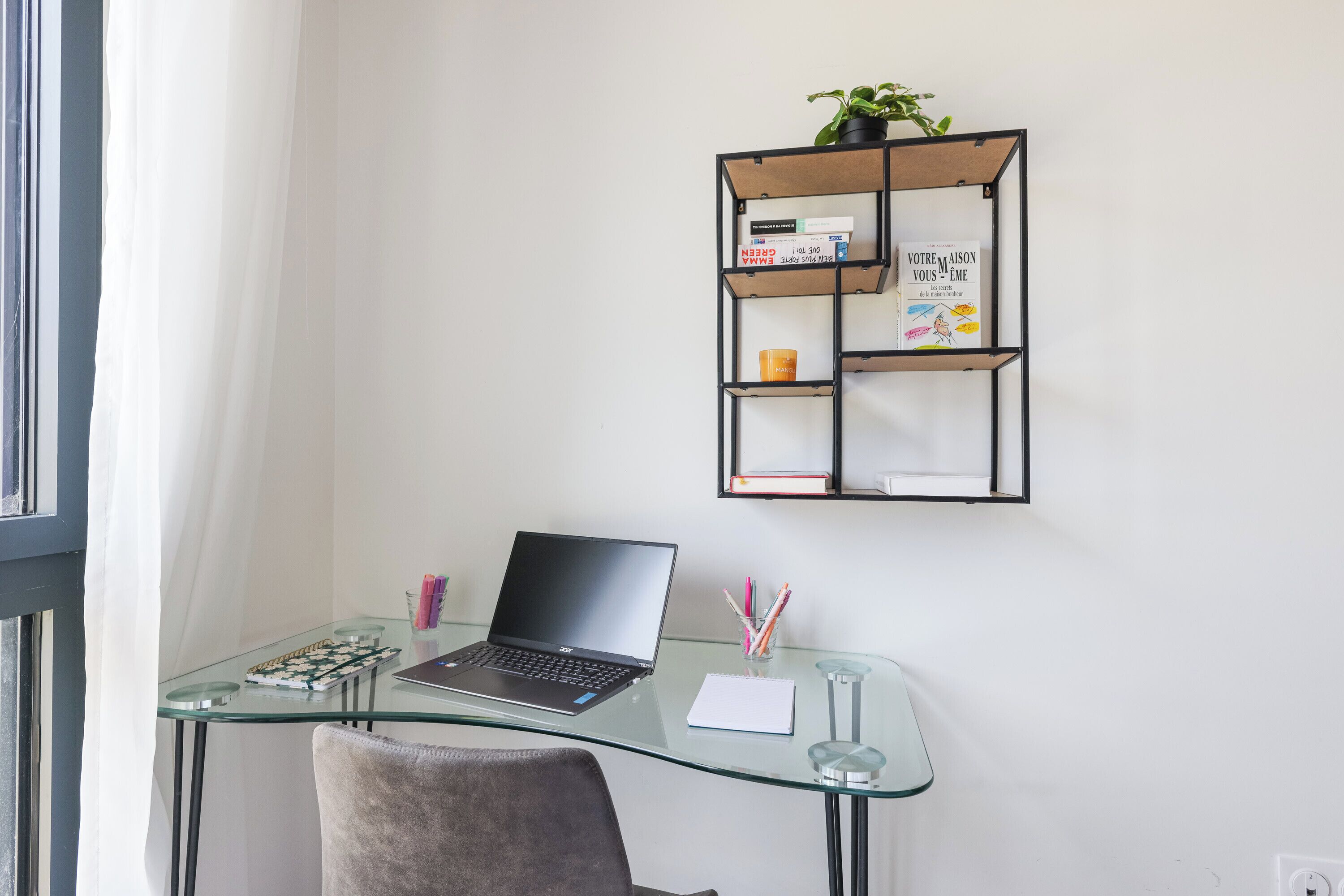 Un escritorio de vidrio con una computadora portátil, una silla, un estante con libros y una planta, y una ventana con cortinas.