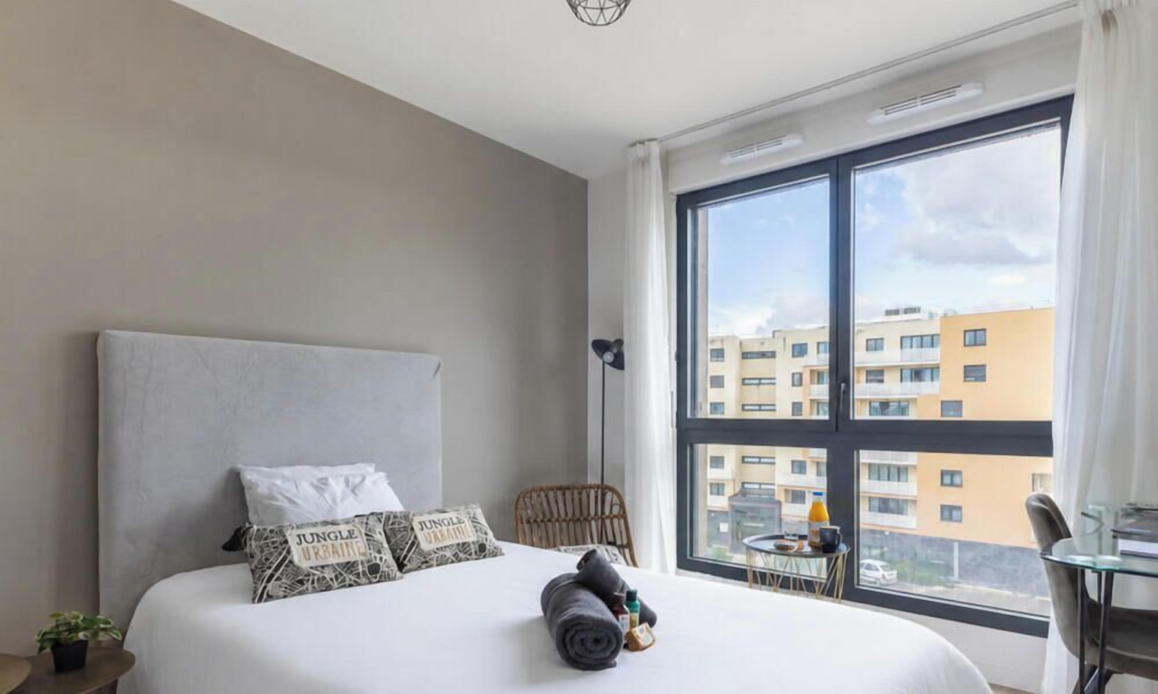 Et hotelværelse med en stor seng, udsigt til bygninger, et lille bord med bakke og en stol.