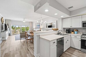 A modern kitchen with white cabinetry, a central island, and a dining area with wooden chairs. - Coastal Retreat at TOPSL - Sleeps 6 (Miramar Beach)