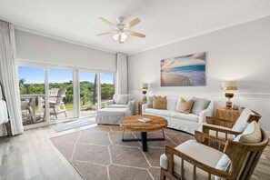 A spacious living room with a large sliding glass door leading to a balcony, a ceiling fan, a round wooden coffee table, and a large painting of a beach scene. - Coastal Retreat at TOPSL - Sleeps 6 (Miramar Beach)