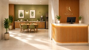 Uma área de recepção de hotel com balcão de madeira, sala de jantar com cadeiras e uma mesa, e obras de arte emolduradas na parede.