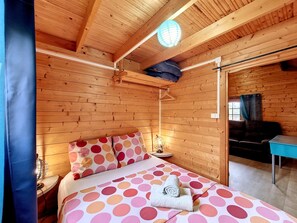 A wooden cabin room with a bed, a hanging light fixture, and a window with curtains. - Joli Cœur - Jacuzzi - Terrasse - Jardin (Saint-Louis)