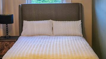 A bed with a headboard, two pillows, a bedside table with a lamp, and a window with a view of greenery.