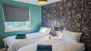 Una habitación con dos camas, una ventana con cortina, una mesita de noche con lámpara y un papel tapiz con motivos florales.