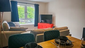 Una sala de estar con un sofá, una mesa de comedor para dos personas, un televisor sobre un mueble rojo y una ventana con cortinas.