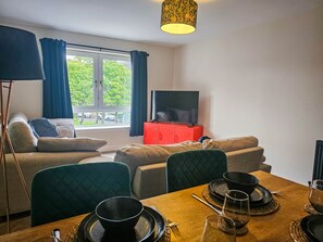 A living room with a sofa, a dining table set for two, a television on a red cabinet, and a window with curtains. - Modern 2-Bed Paisley: RAH · Airport · Free Parking (Paisley)