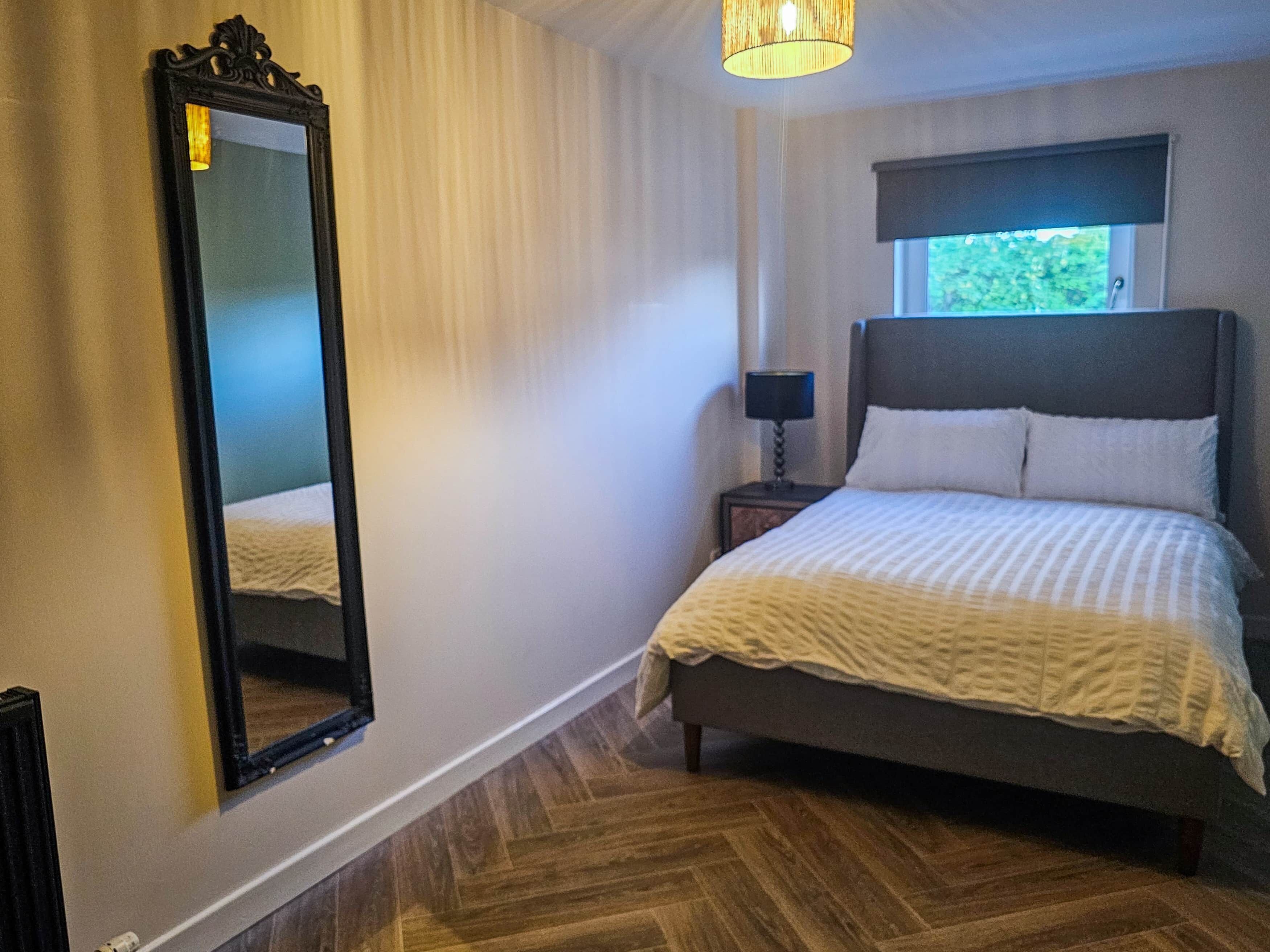 A bedroom with a bed, a nightstand, a lamp, a window, and a mirror.