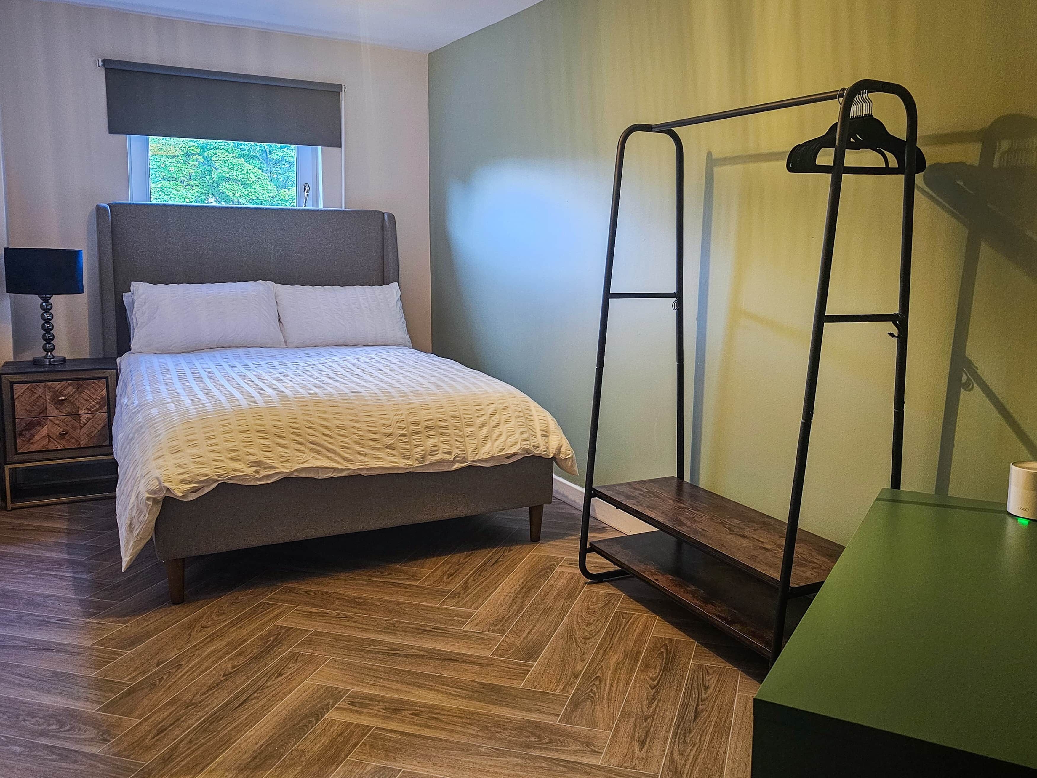 A neatly arranged bedroom with a bed, bedside table, lamp, and a standing fan.