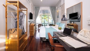 Une chambre d’hôtel avec un lit, un bureau avec un ordinateur portable, une télévision, une étagère en bois avec un sac, et une fenêtre donnant sur un paysage.