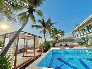 A swimming pool with lounge chairs, a covered seating area, and palm trees.