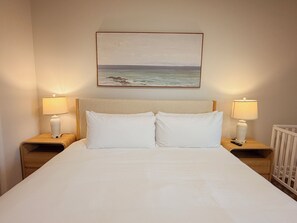 Uma cama bem arrumada com lençóis brancos, duas mesas de cabeceira com abajures e um quadro de uma paisagem costeira sobre a cabeceira.