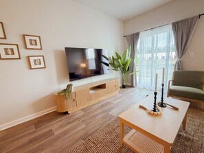 En stue med et fladskærms-tv, et tv-møbel i træ, en grøn plante, et stort vindue med gardiner og en grøn lænestol.