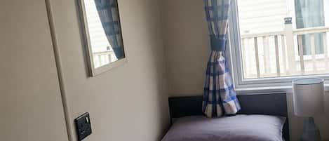 A single bed with a headboard, a bedside table with a lamp, a power outlet, a window with curtains, and a mirror on the wall.