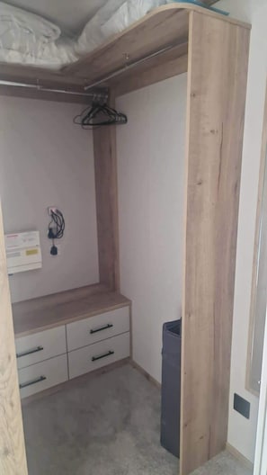 Une pièce comprenant une armoire en bois, un petit bureau avec des tiroirs, un portant à vêtements et une poubelle.