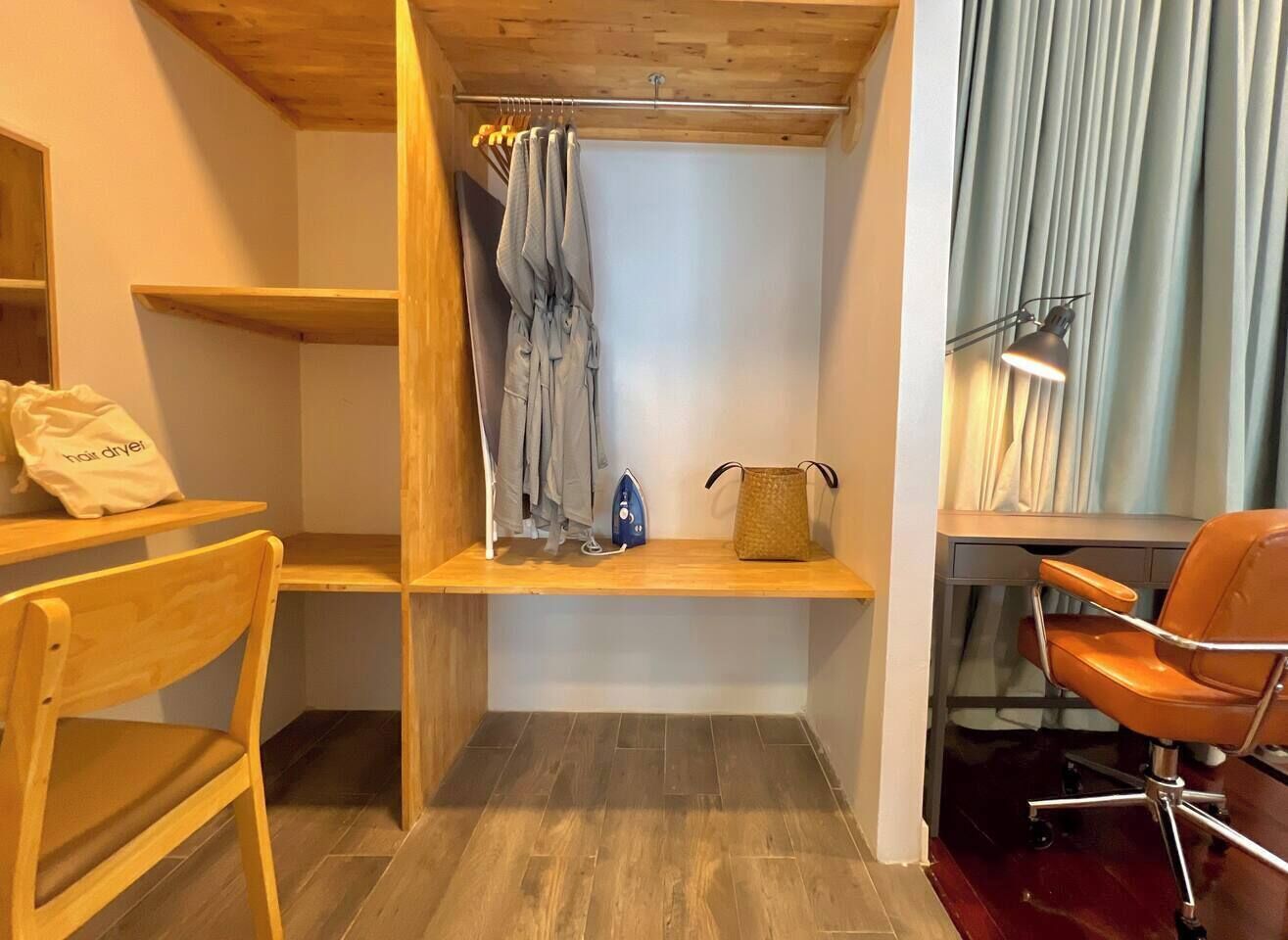 Une pièce de taille modeste, comprenant un bureau en bois, une chaise, un vêtement suspendu, une petite étagère et une lampe.