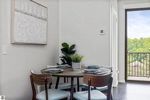 Uma sala de jantar com uma mesa redonda, seis cadeiras, um vaso de planta e vista através de uma porta de vidro.