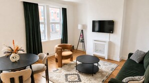 Ein Wohnzimmer mit rundem Tisch, zwei Stühlen, einer Couch, einem Fernseher und einem Fenster mit Vorhängen.