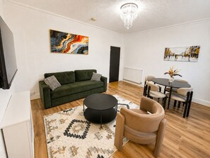 Ein modernes Wohnzimmer mit einem grünen Sofa, einem runden schwarzen Couchtisch, einem beigen Sessel, einem Esstisch mit Stühlen und zwei Kunstwerken an den Wänden.