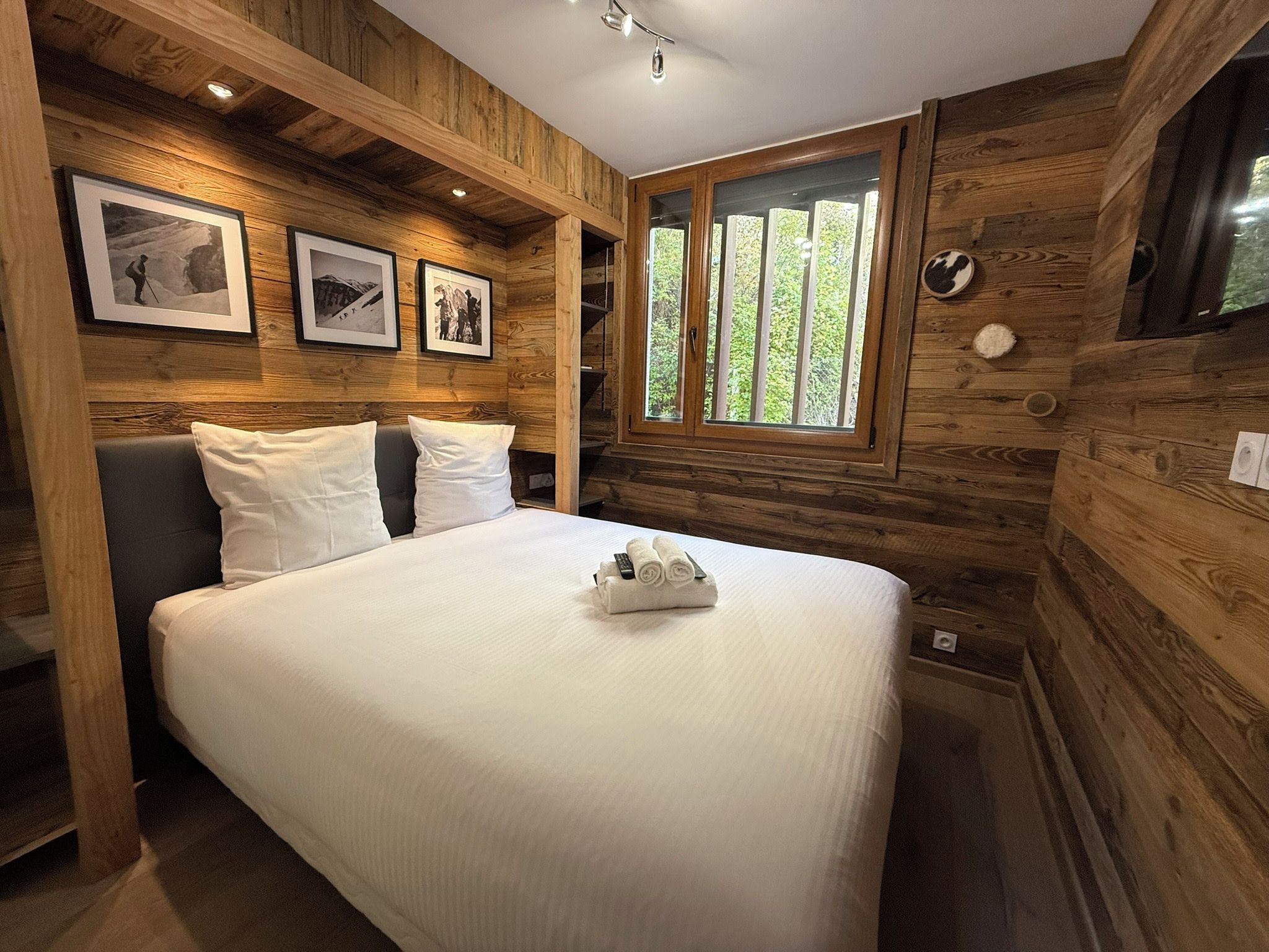 A wooden-paneled room with a bed, framed pictures on the wall, and a window with a view of greenery.