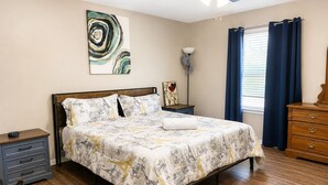 A bedroom with a bed, a nightstand, a dresser, a lamp, a painting, and a window with curtains. - Private Pool, Near NRG Stadium & Texas Medical & Downtown (Houston)
