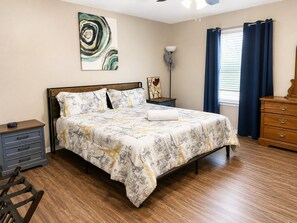 Een slaapkamer met een bed, nachtkastje, kapstok, lamp, schilderij en raam met gordijnen.