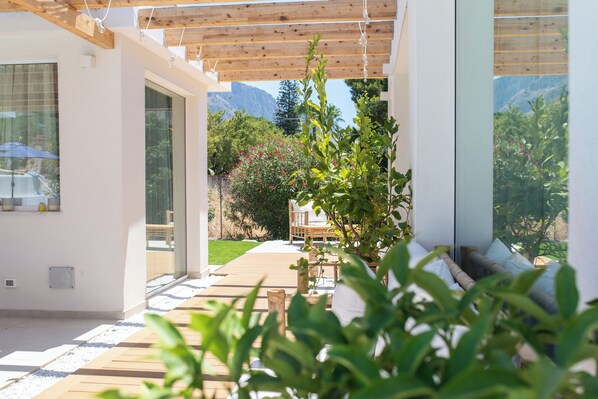A modern outdoor patio with wooden decking, a glass door leading to a garden, and a view of a mountain.