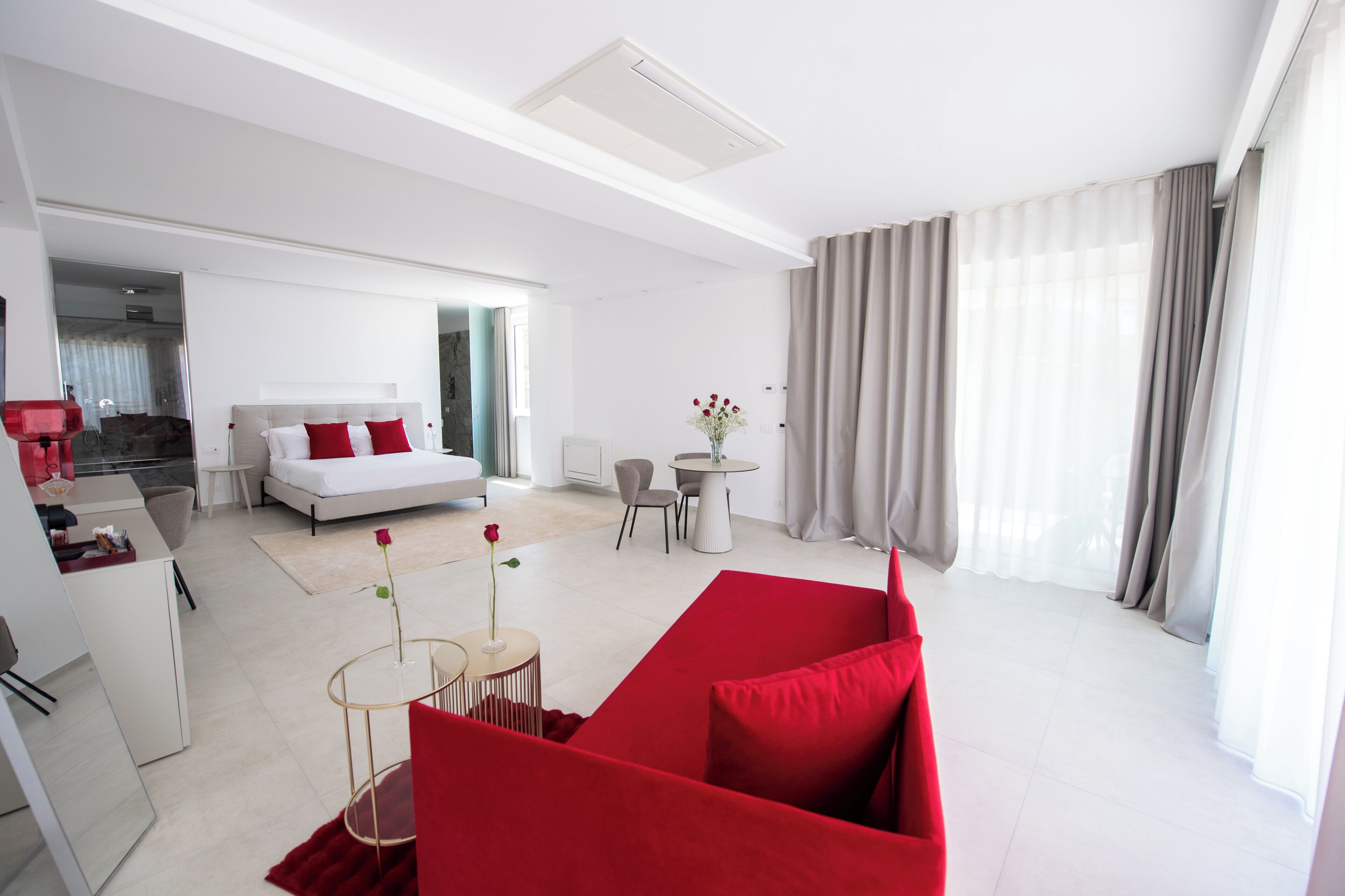 A modern hotel room with a bed, a red sofa, a small table with a vase, and a view of the city.