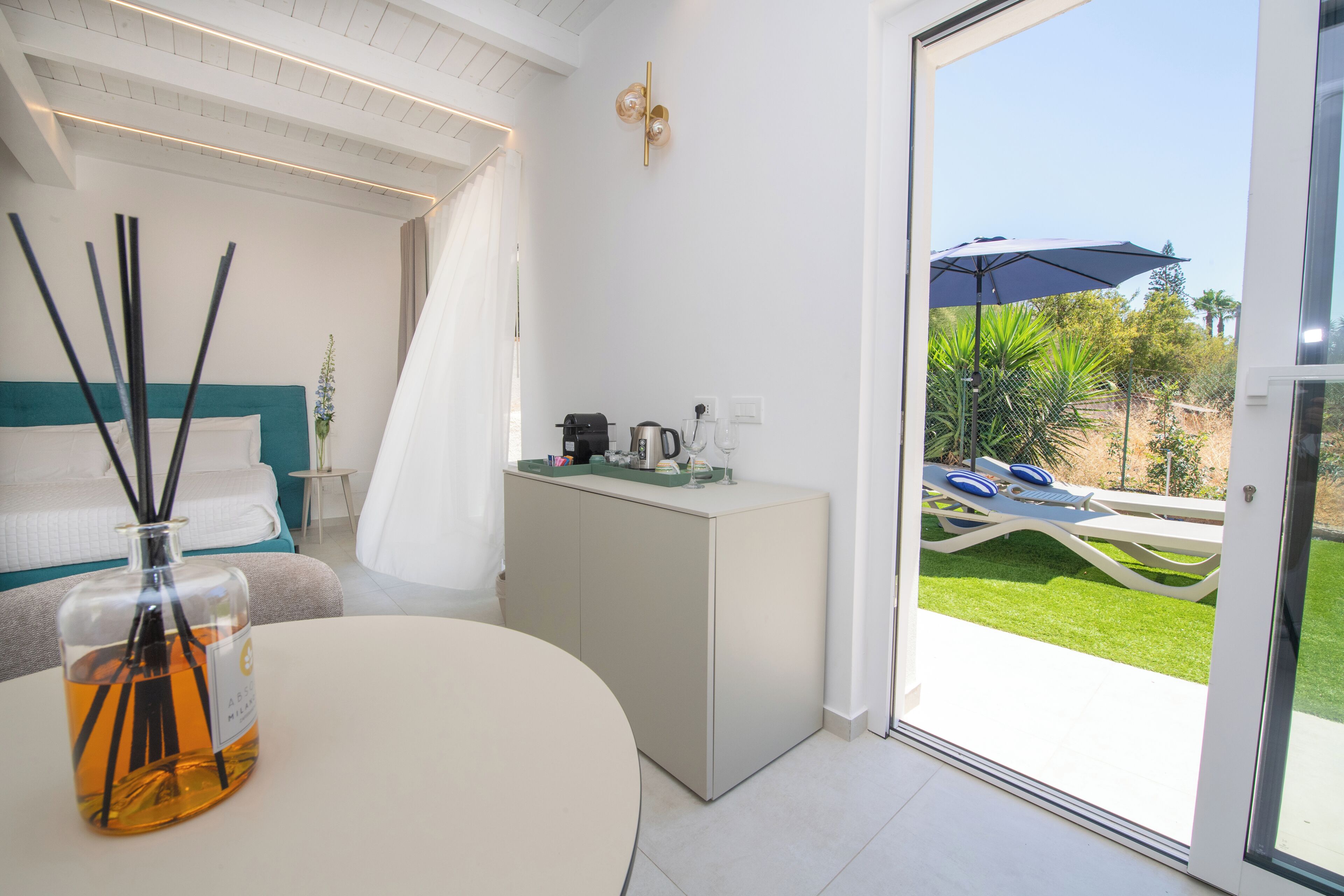 Een moderne slaapkamer met een bed, nachtkastje, klein kastje en uitzicht op een terras met tafel en stoelen.