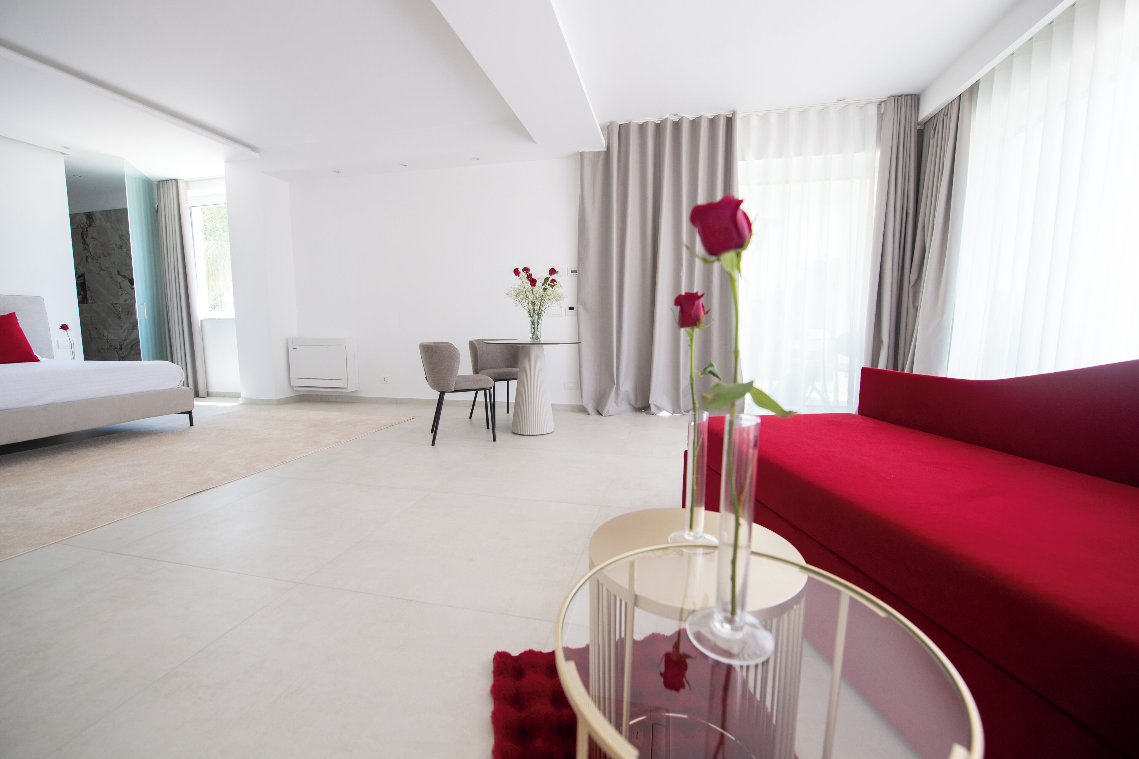 A modern hotel room with a bed, a red sofa, a glass coffee table with a vase of flowers, a dining area with a table and chairs, and a view of the city through large windows.