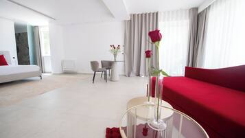 A modern hotel room with a bed, a red sofa, a glass coffee table with a vase of flowers, a dining area with a table and chairs, and a view of the city through large windows.