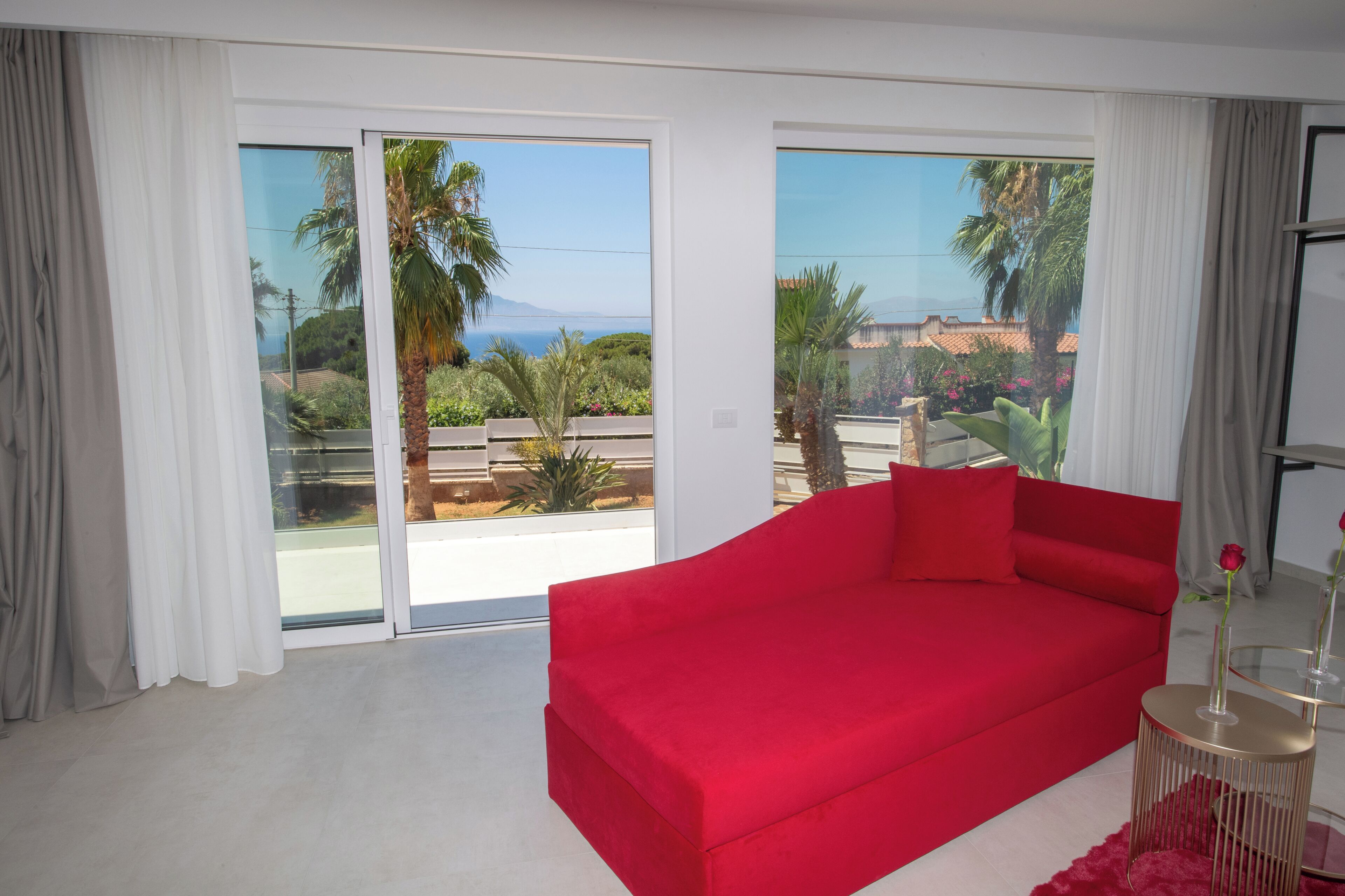 Een rode bank in een kamer met grote schuifdeuren die uitkomen op een balkon met uitzicht op palmbomen en een ver landschap.