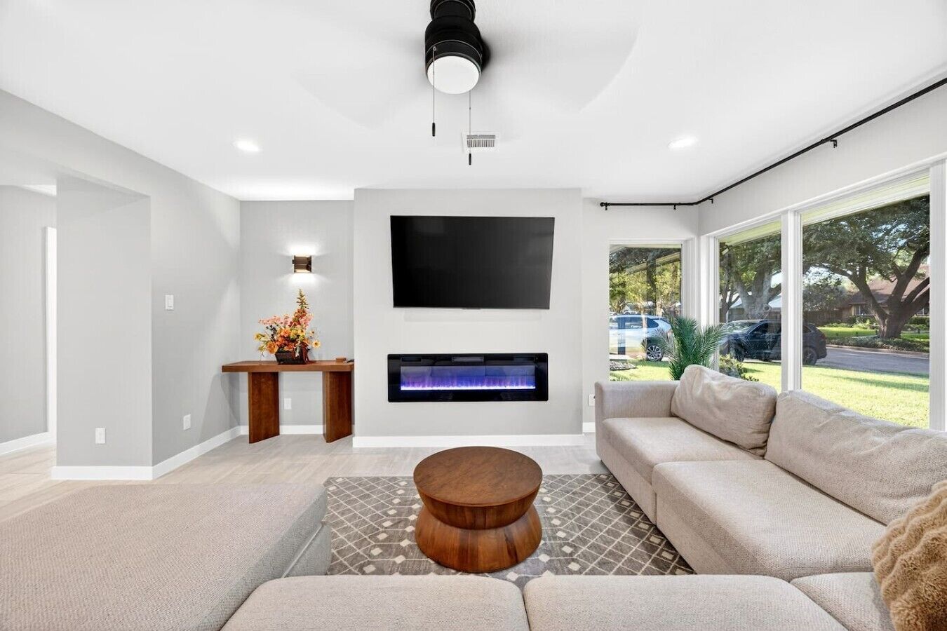 Una sala de estar moderna con un sofá, una mesa de centro, una chimenea, un televisor montado en la pared y un ventanal con vistas al exterior.